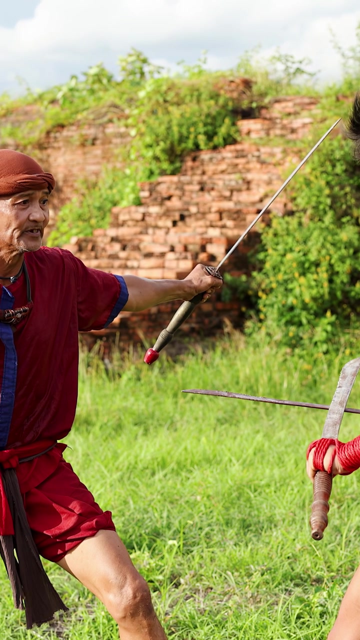 Due uomini in abiti tradizionali rossi praticano arti marziali con spade tra antiche rovine.