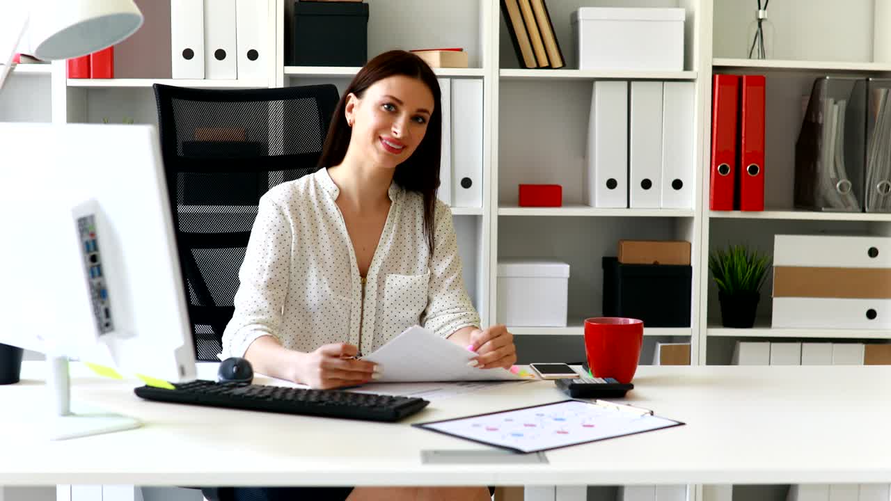 Empresária de blusa branca sentada à mesa e olhando para a câmera.