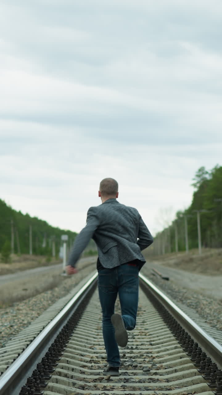 A close view of an aged man running on a railway track, looking back and then forward, he is wearing a grey suit, jeans, and canvas shoes, with trees, electric poles, and two yellow poles