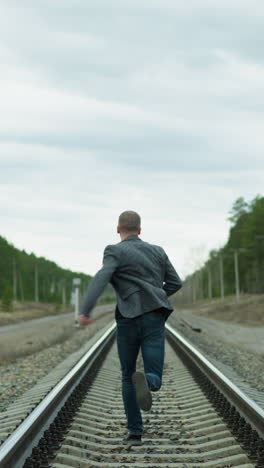 una vista cercana de un hombre anciano corriendo por una vía ferroviaria, mirando hacia atrás y luego hacia adelante, está vestido con un traje gris, vaqueros y zapatos de lona, con árboles, postes eléctricos y dos postes amarillos