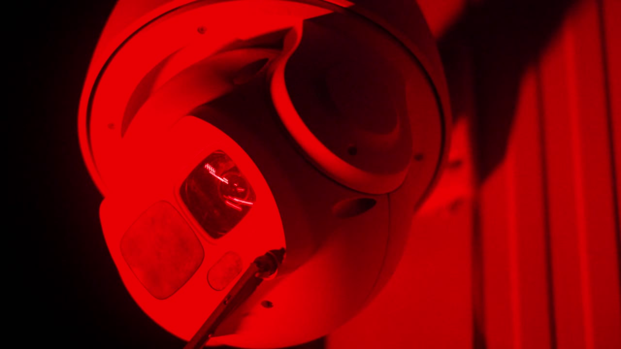 Fixed CCTV security camera mounted on a building corner in darkness, lit only by red and blue emergency service lights. Implied police response, surveillance, public safety, urban crime monitoring