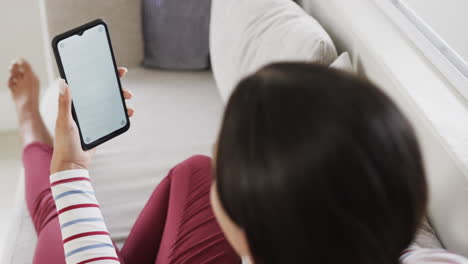 Biracial-woman-sitting-on-sofa-using-smartphone-with-copy-space-on-screen,-slow-motion