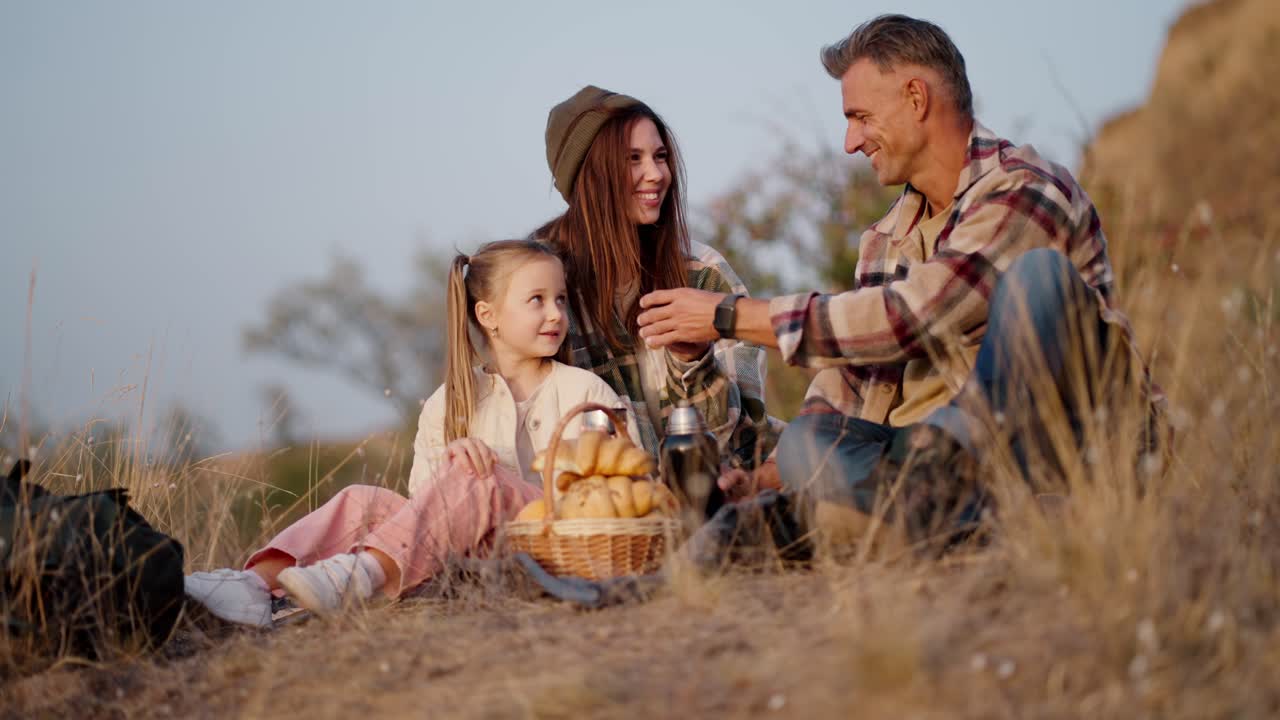 Side shot of a happy middle-aged man with gray hair in a checkered shirt pours tea for his wife and little daughter during their picnic outside the city in the summer evening