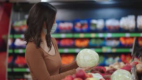 una mujer con una máscara en un supermercado elige frutas y verduras sosteniendo repollo en sus manos en una tienda