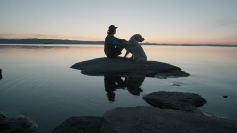 Mujer-Acariciando-A-Un-Perro-En-Una-Roca-En-El-Lago-Al-Atardecer