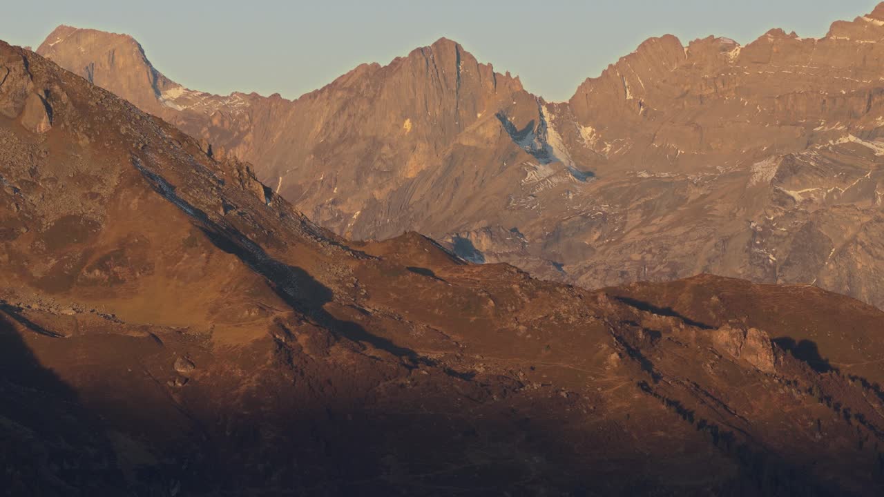 Aerial drone footage captures Swiss alpine peaks during sunrise. Soft golden light enhances the textures of the mountains. Calm parallax motion reveals layered mountain ranges in cinematic style