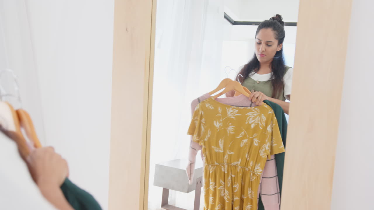 Choosing between outfits, asian woman holding dress and sweater in front of mirror, at home