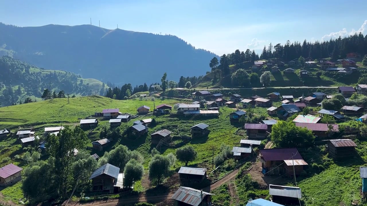 Vista panorâmica de paisagem montanhosa de verão e vila tranquila, com floresta de pinheiros em encosta verde, em terreno natural com cabanas espalhadas, vegetação exuberante, prado ensolarado, para uma vibrante experiência de viagem aérea remota em Savsat