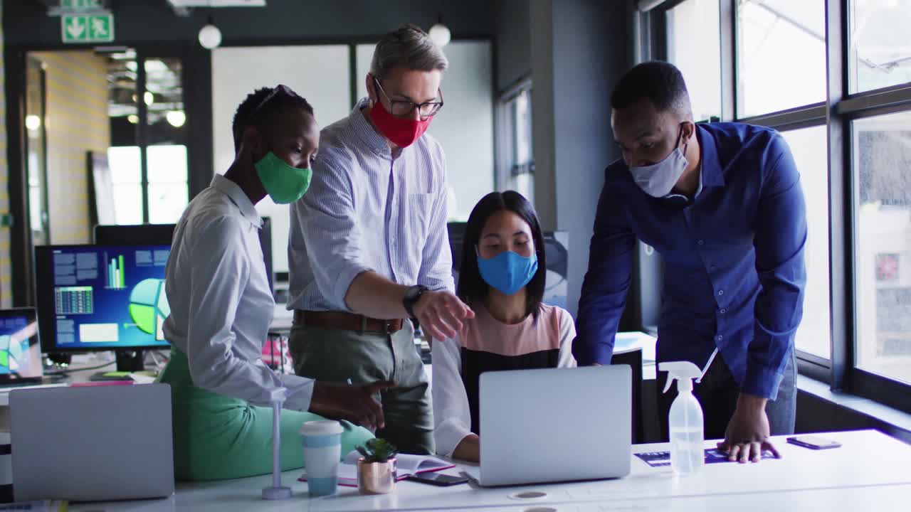 Office colleagues wearing face masks discussing over laptop at modern office