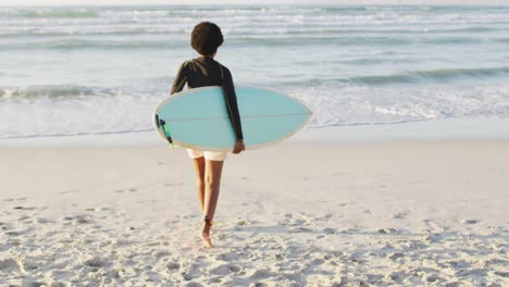 Afroamerikanische-Frau,-Die-Mit-Surfbrett-Am-Sonnigen-Strand-Spaziert