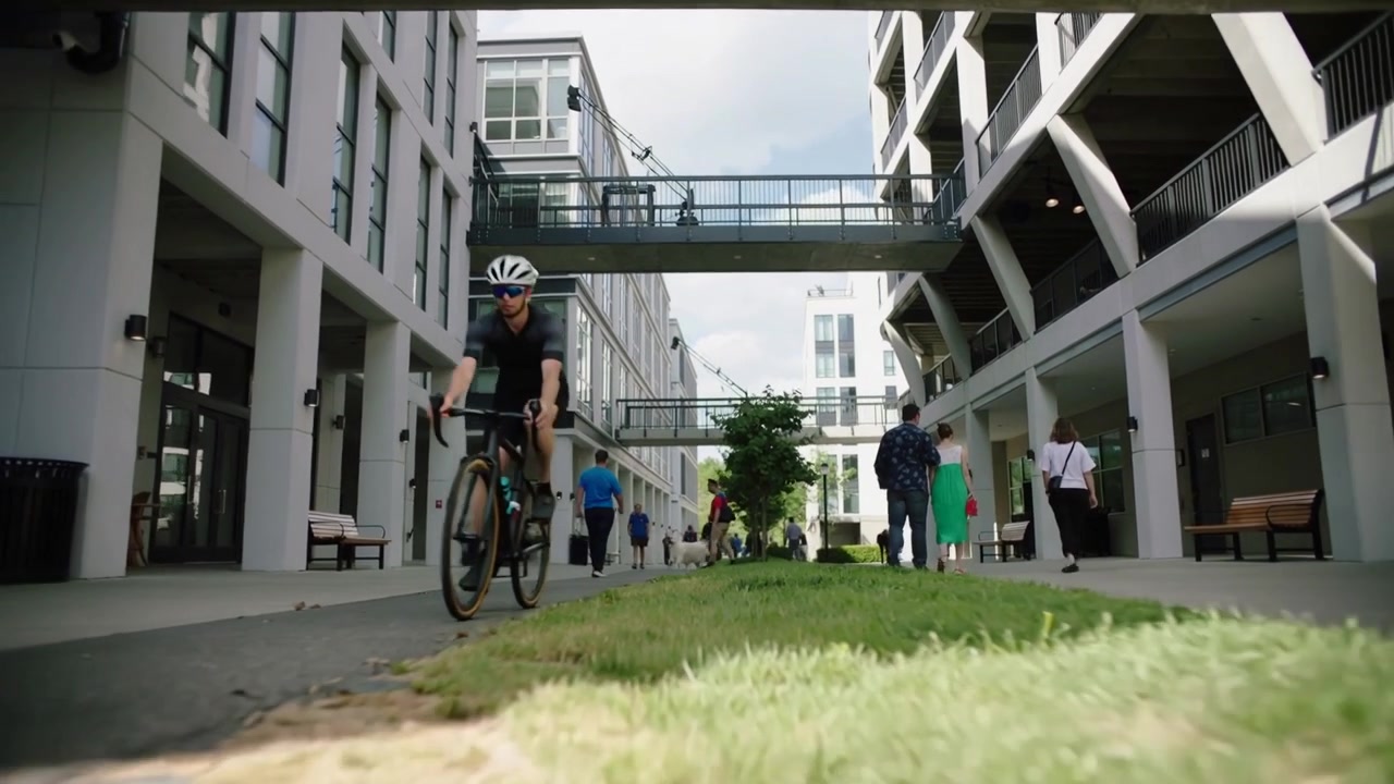 En cyklist nærmer sig og passerer kameraet gennem en gårdsplads' midterrabat, iført hjelm og cykeltøj på vej til arbejde.