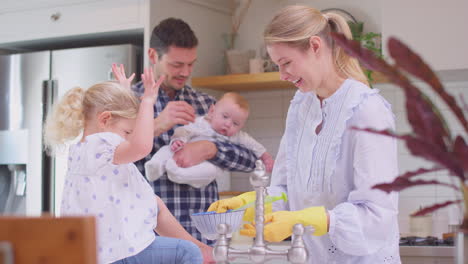 Vater-Und-Kleiner-Sohn-Schauen-Zu,-Wie-Die-Tochter-Auf-Der-Theke-Sitzt-Und-Der-Mutter-Beim-Abwaschen-Des-Geschirrs-Im-Waschbecken-Hilft-–-Aufgenommen-In-Zeitlupe