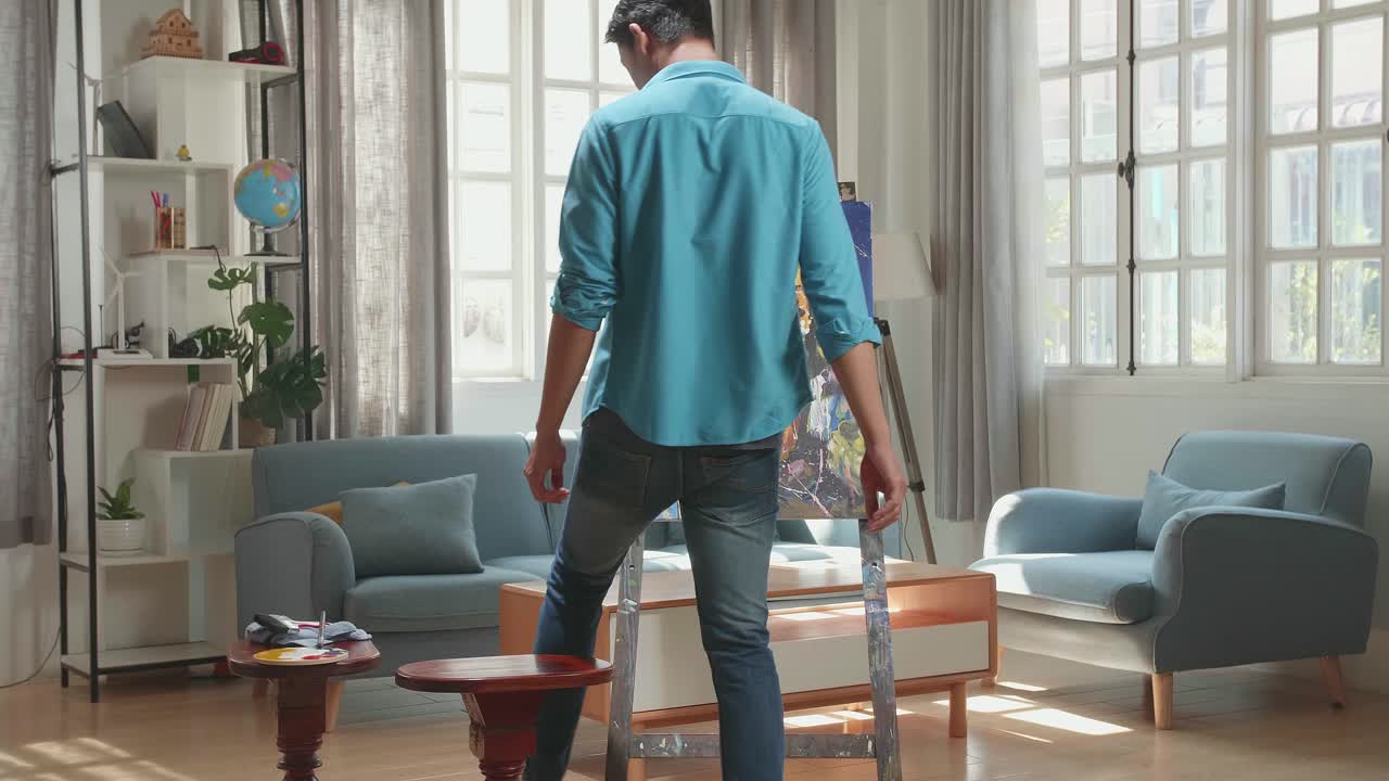 vista trasera de un artista asiático hombre que viene a sentarse en una silla de madera redonda sin respaldo y sosteniendo un pincel de color mixto antes de pintar a una chica en el lienzo