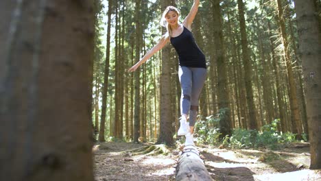 una joven vestida con ropa deportiva camina juguetonamente sobre un árbol caído usando sus manos para mantener el equilibrio y patea su pierna antes de saltar hacia abajo.