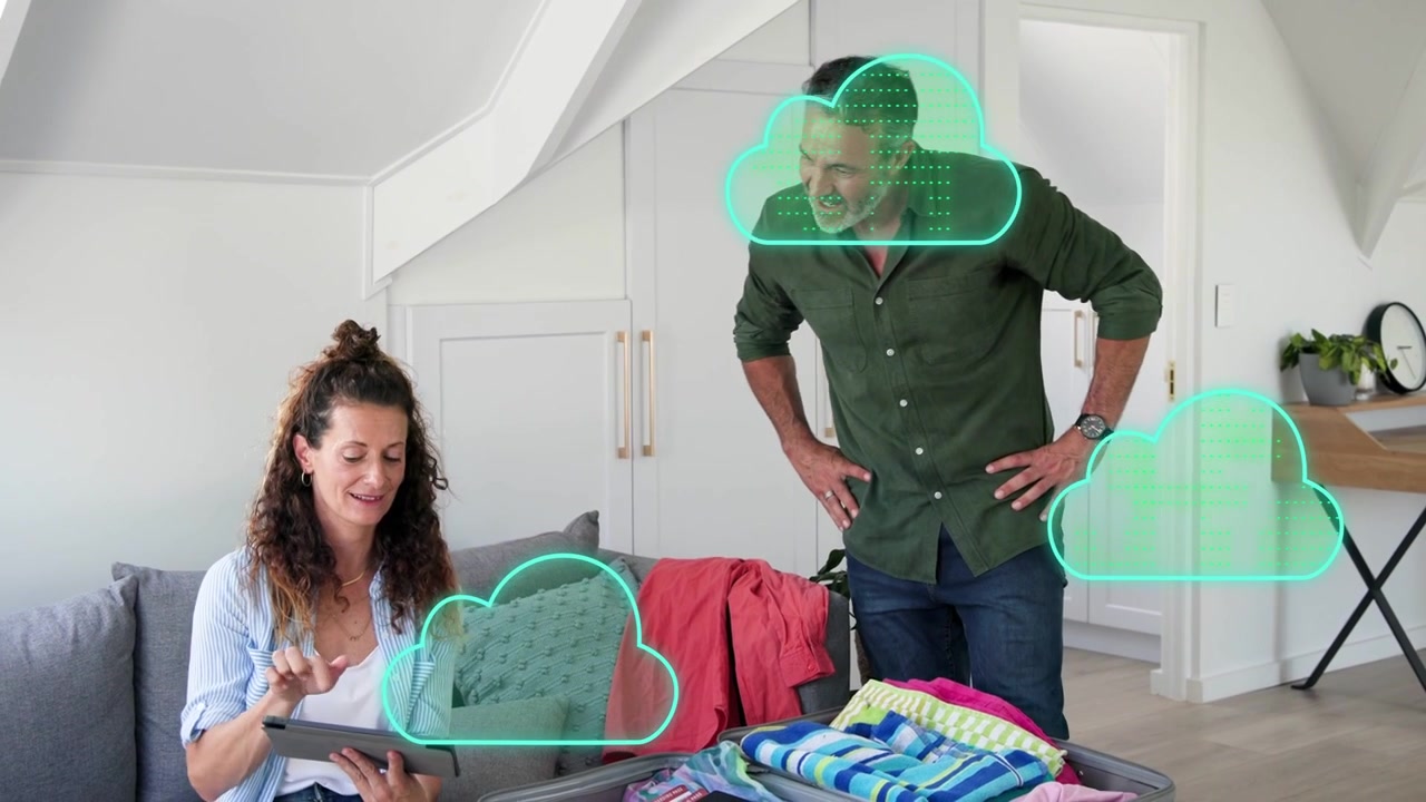 Coppia che prepara la valigia in un loft, donna che tocca il tablet creando nuvole al neon che collegano articoli da viaggio.