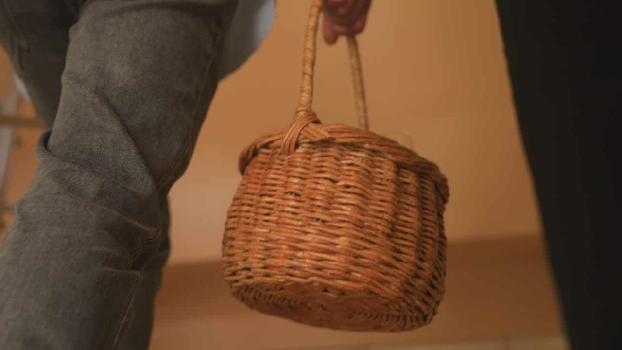 Casal carregando uma cesta de piquenique de vime pelas escadas, clima noturno, jeans e roupas escuras, garrafa de vinho e taças visíveis na cesta, iluminação interna quente, subida suave, chegada para um encontro noturno íntimo, lar