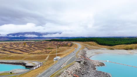aerial view of a vehicle traveling a lakeside road with turquoise water and mountainous backdrop under cloudy skies