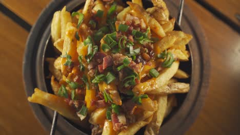 una toma estacionaria de arriba hacia abajo de un tazón lleno de tocino y papas fritas cubierto con queso cheddar y cubierto con cebolletas