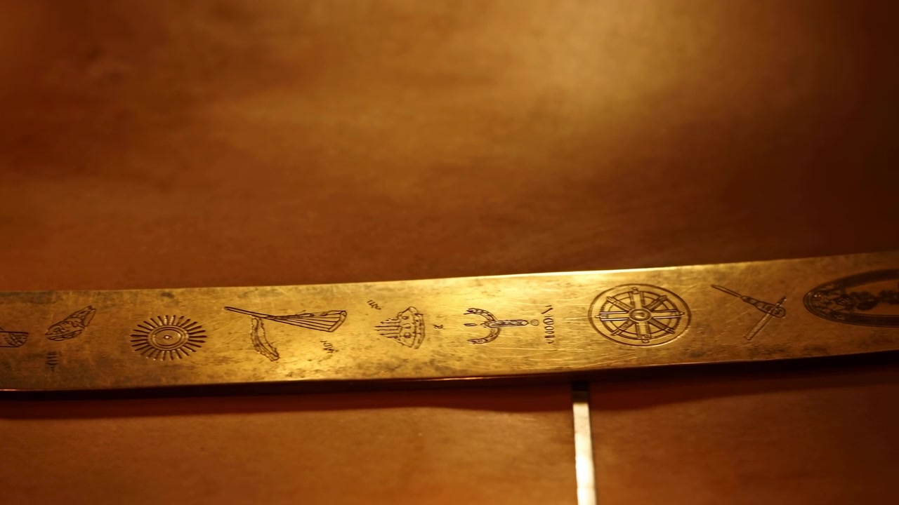 Primo piano di una striscia di ottone incisa che poggia lungo il bordo inferiore, illuminata dalla fiamma di una candela con un bagliore caldo, riflessi tenui, sfondo scuro e texture sottile.