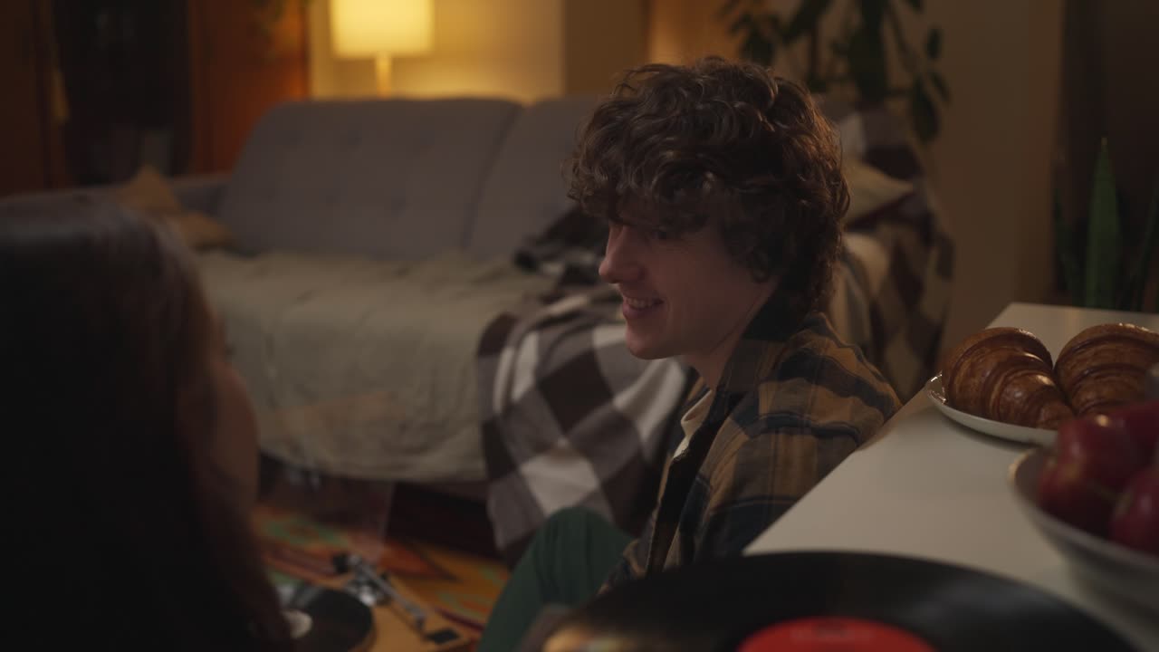 A young couple enjoys conversation in a cozy living room with croissants and records