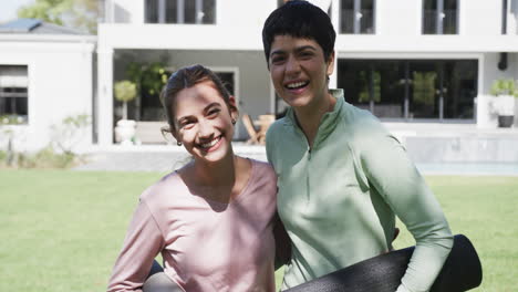 Feliz-Pareja-De-Lesbianas-Caucásicas-Paradas-Con-Estera-De-Yoga-Y-Sonriendo-En-Un-Jardín-Soleado,-Cámara-Lenta