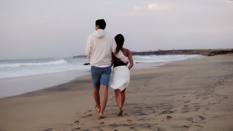 Relaxed-young-couple-on-beach-walking-in-love-man-holding-his-girl-by-from-back-.-Woman-and-man-relaxing-on-travel-vacation-holidays,-man-wearing-casual-clothes,-woman-white-towel-on-belly.-Slow-motion.-Rare-view