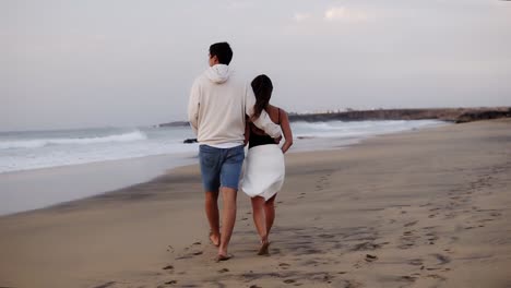 relaxed young couple on beach walking in love man holding his girl by from back . woman and man relaxing on travel vacation holidays, man wearing casual clothes, woman white towel on belly. slow motion. rare view