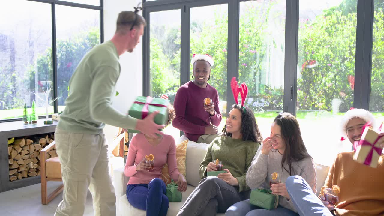 Amid eager diverse friends, mid adult white man entering home carrying wrapped gifts, handing them