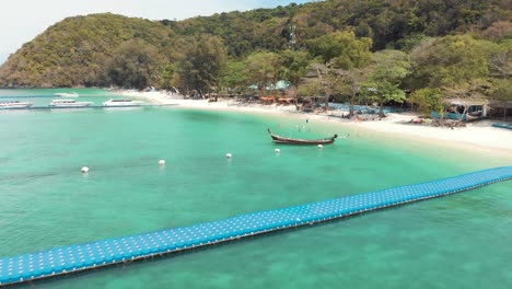 touristic banana beach in koh hey with long-tail fishing boat moored near emerald shore, thailand - aerial low angle fly-backwards shot