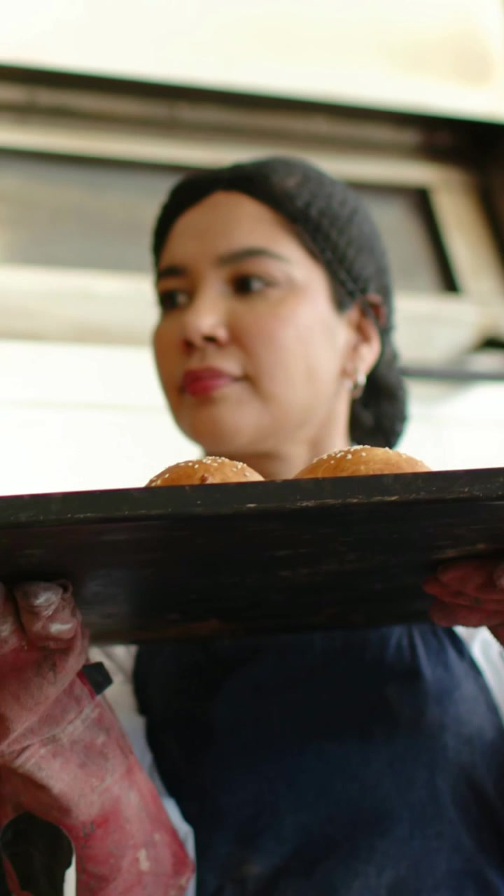 Vertical video: Oven opening bakery chef in coat pulling tray checking placing buns on rack cooling