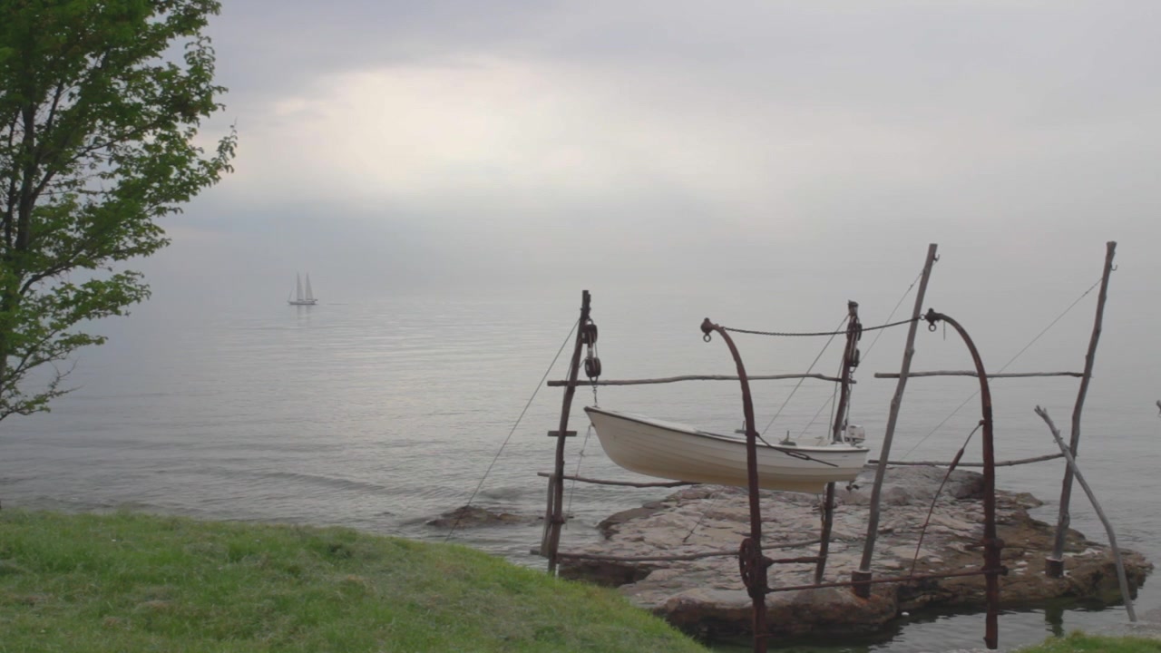 Una barca a vela bianca a due alberi naviga sul mare lungo la costa croata in una tranquilla e nuvolosa giornata estiva, in un'inquadratura ampia. Un'altra barca è ormeggiata a una struttura di molo in legno e roccia in primo piano, tra campi verdi e alberi.