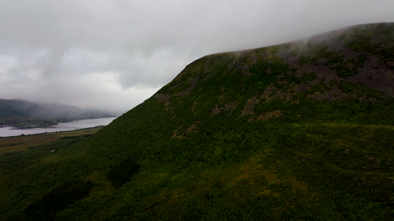 Lush green mountain slope disappearing into low clouds. The town of Sortland and the fjord are visible in the distance under a moody grey sky in Northern Norway. Aerial forward