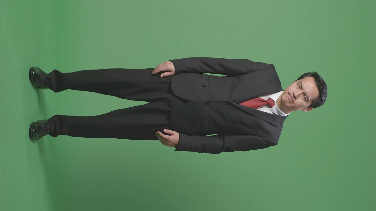 아시아 사업가의 몸이 가득 차 있으며, 만족하지 않고 녹색 스크린 앞에 서서 머리를 흔들고 있습니다.