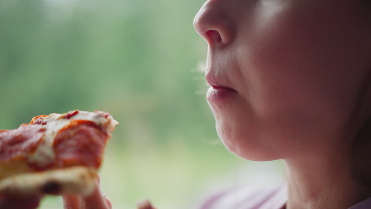 Close up of young girl enjoying toasted bread near window with natural soft daylight, blur greenery visible outside, focus on hand holding bread and lips taking bite