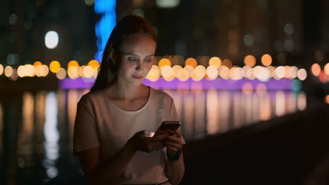 at night a young girl sits on the grass in a big city and looks into the smartphone screen writes a message and maintains her personal blog.