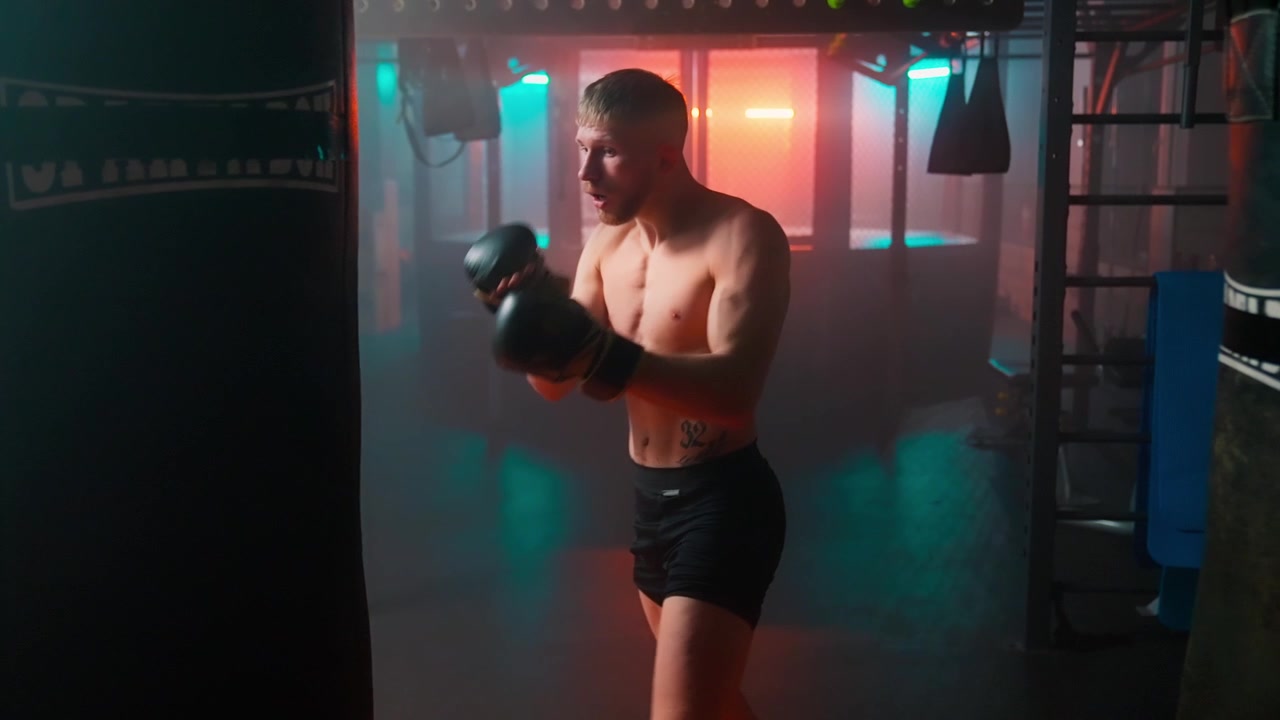 Shirtless Male Boxer Training with Punching Bag in Dark Gym with Neon Lights