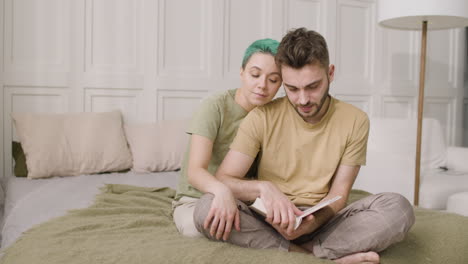 joven sentado en la cama y sosteniendo un libro mientras su novia lo besa y lo abraza