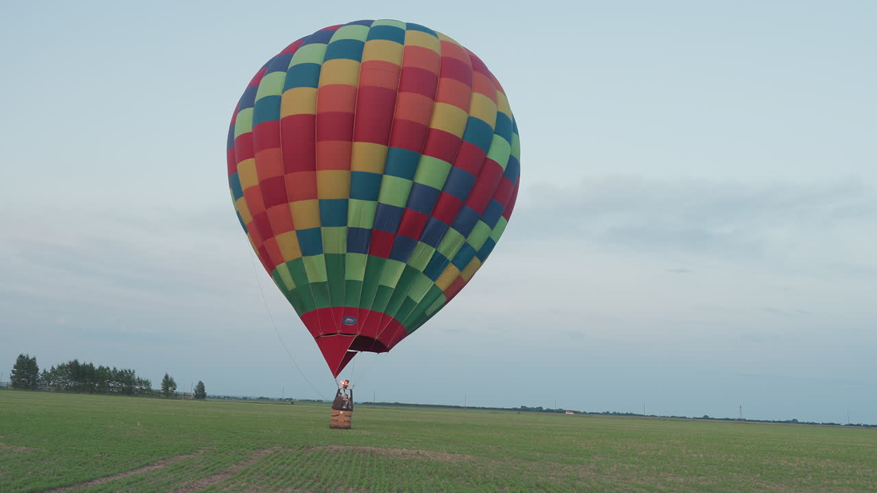 rear view passenger riding multicolor balloon gliding low over green farmland at dusk with flame flickering under basket against pastel sky