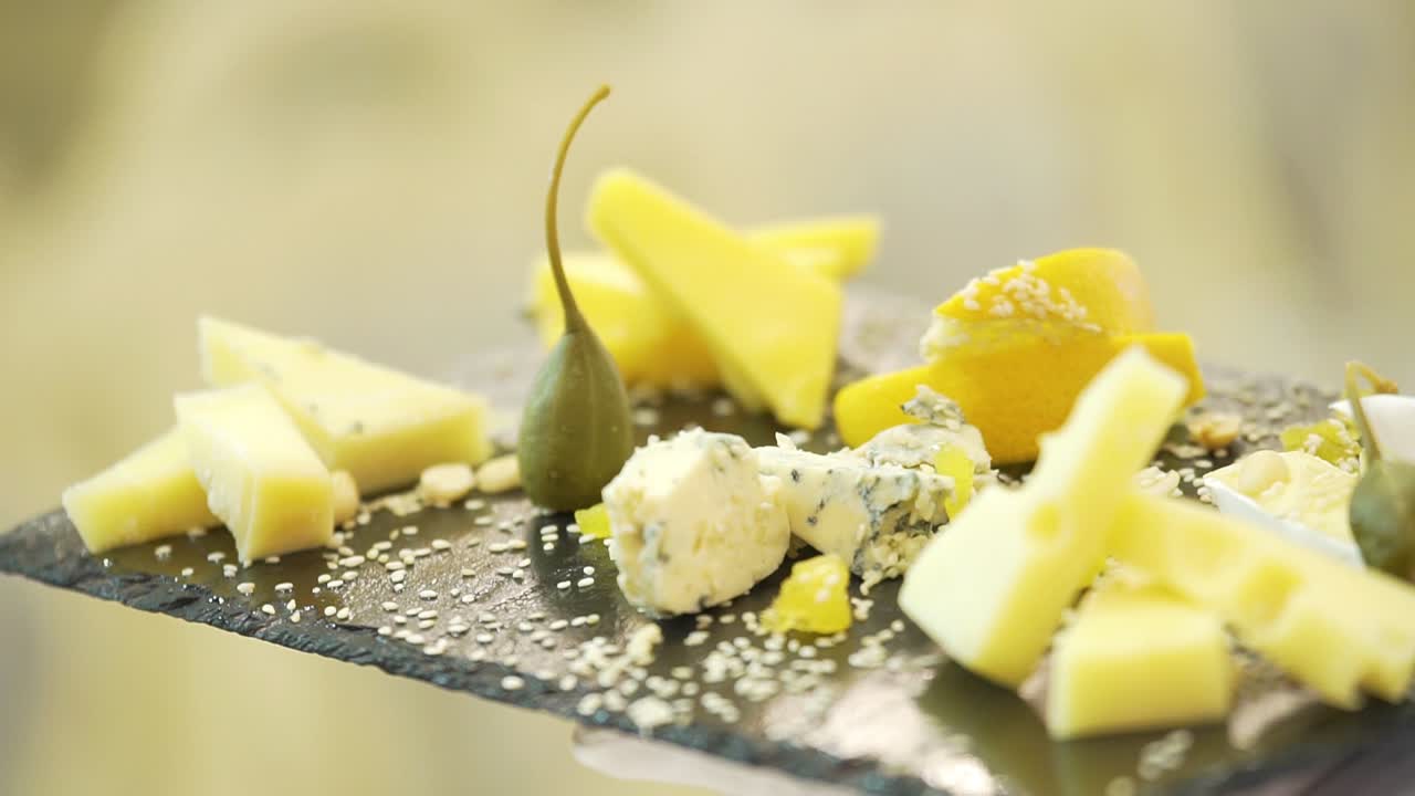 A waiter in white gloves holds a black wooden board with pieces of various hard cheese sprinkled with sesame seeds and gently puts green fruit near it. Close-up.