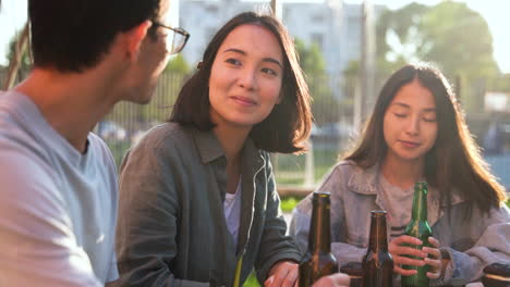 group of three japanese friends with beers talking together while sitting at table outdoors in a sunny day