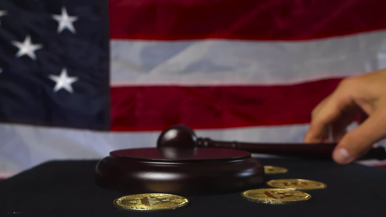 Slow motion video of a justice court hammer or gaol hitting a wooden board while golden shiny bitcoin money cryptocurrency coins are on the side, flying and bouncing from the impact. American flag