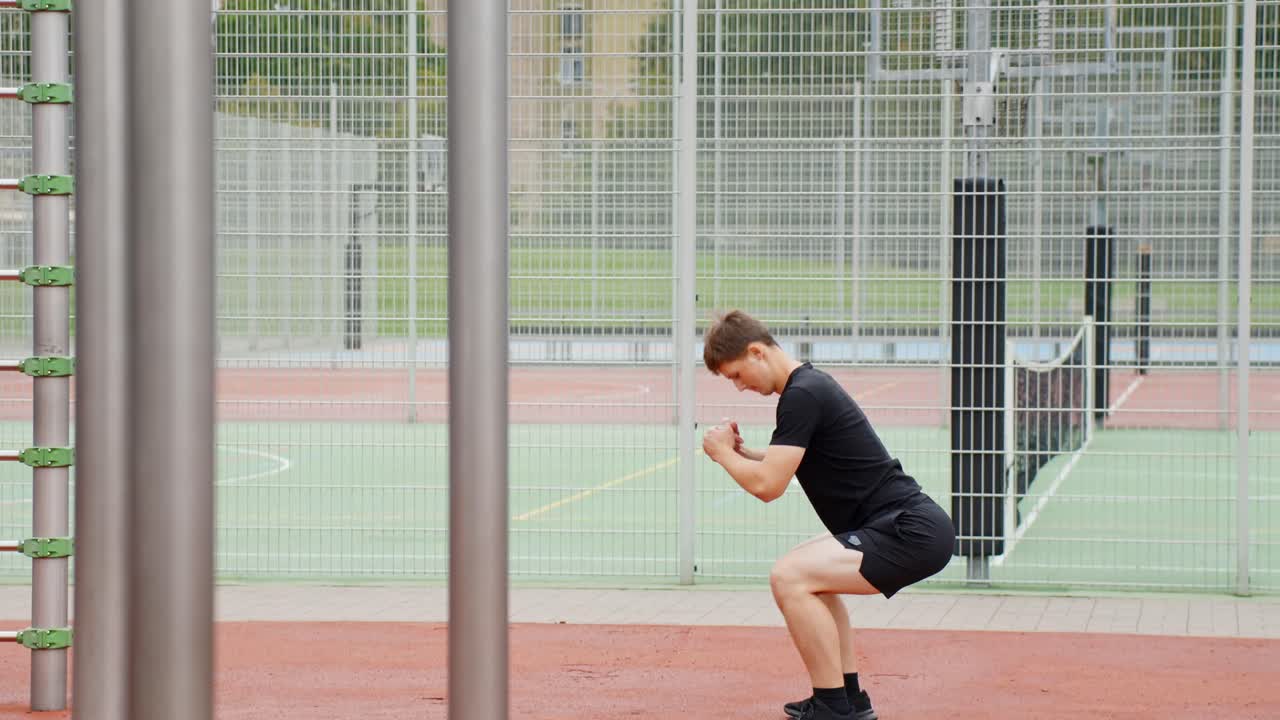 Inquadratura fissa di un giovane che fa squat in un'area sportiva all'aperto, mostrato di profilo, concentrato e impegnato