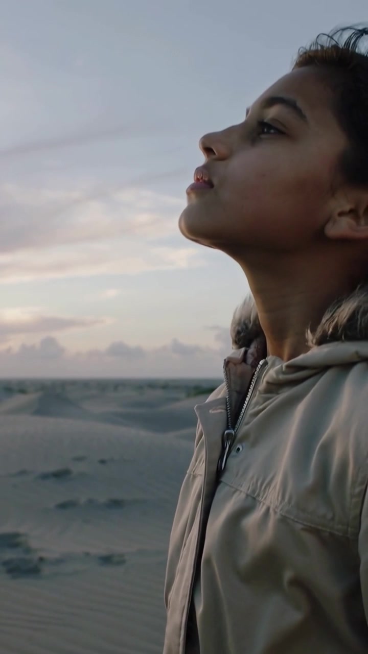 Video verticale: Telecamera in ascesa che mostra un adolescente con cappuccio di pelliccia che guarda in alto sulle dune all'alba, con spazio per il testo.