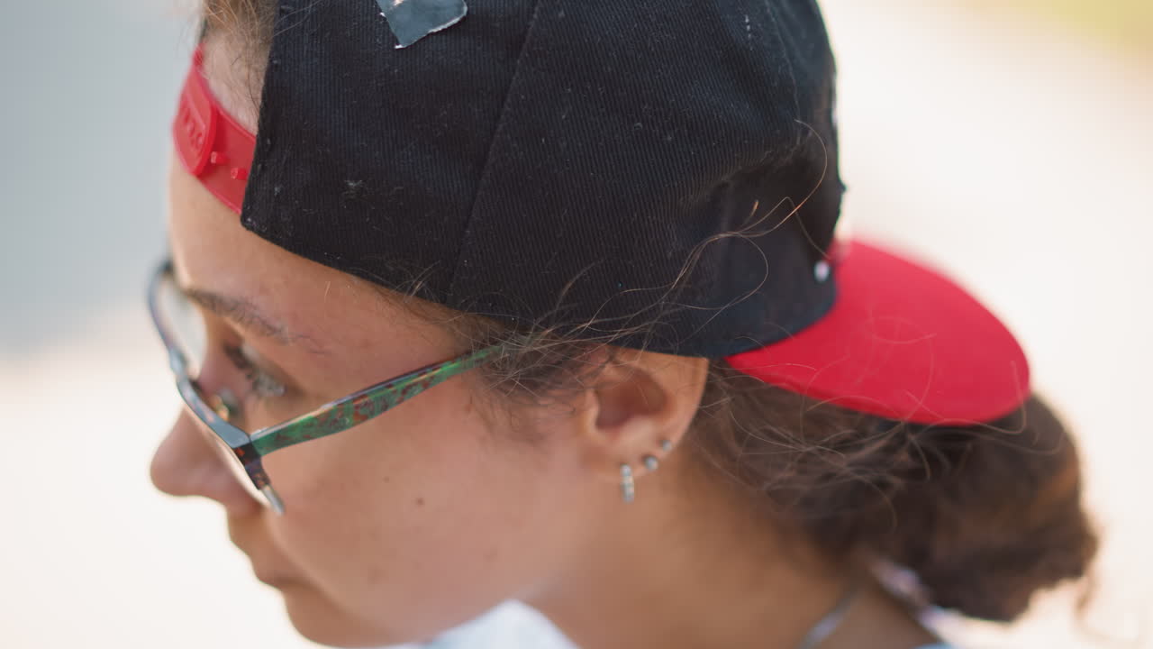 Joven con gorra y auriculares preparándose para correr en una calle urbana soleada, ajustándose los auriculares y mirando el reloj, con gafas y gorra hacia atrás; los primeros planos enfocados capturan la rutina casual