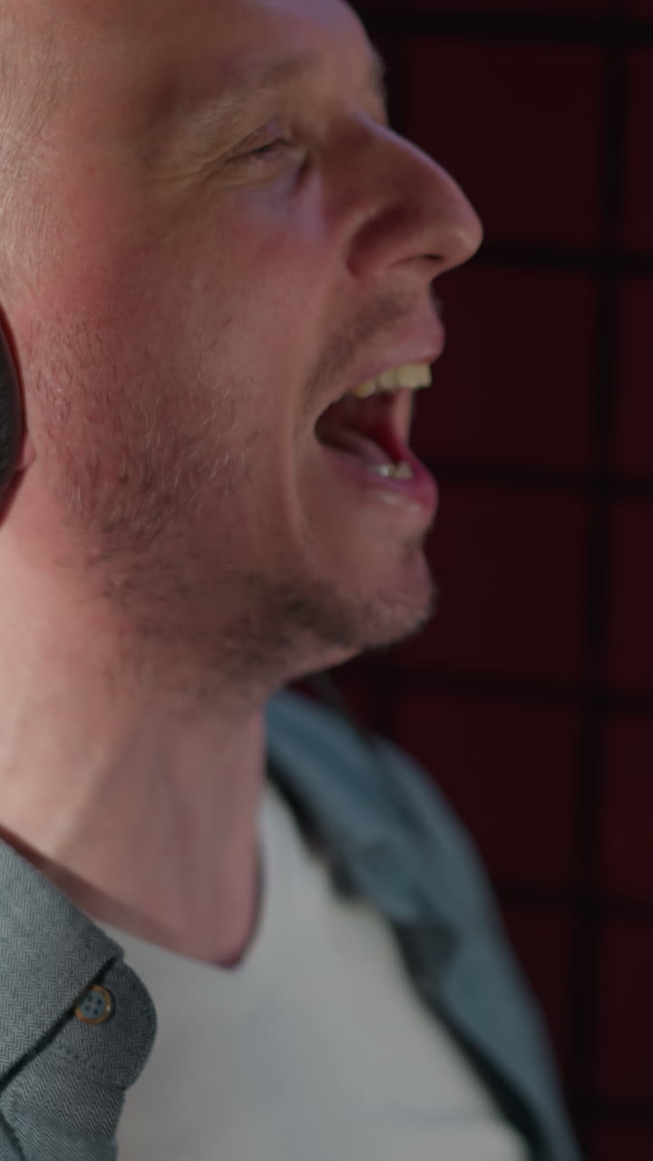 una toma lateral de primer plano de un hombre cantando apasionadamente con la boca abierta y los ojos cerrados. está usando auriculares negros y está ambientado contra un fondo rojo en un estudio de grabación