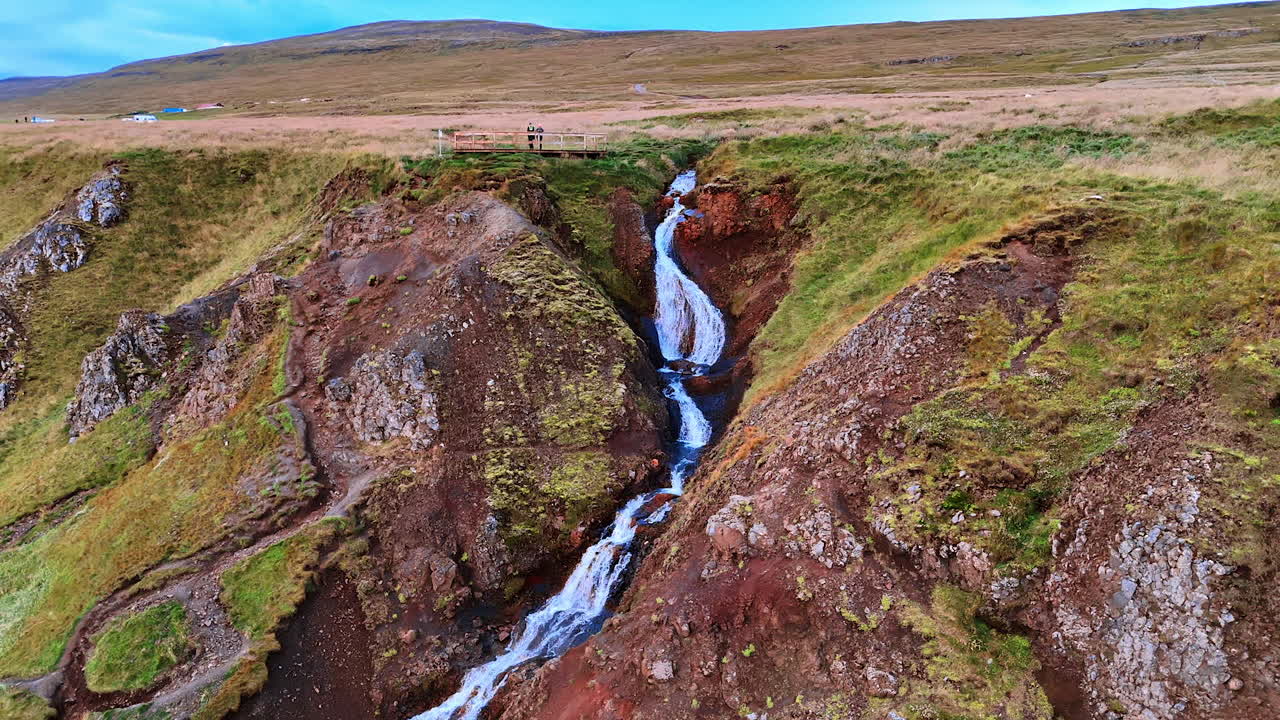 Two people stand on the observation deck located on the mountain. Tourists watch the waterfall flows by the rocks. Top view. Iceland landmarks.