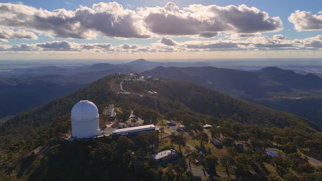 Aerial drone footage glides over a mountaintop observatory, capturing a large telescope dome, forested hills, and dramatic sunset lighting with scattered clouds