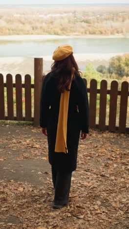 rear view of a tourist in a yellow beret and black coat walking towards a wooden fence, resting her hand on it, enjoying the scenic view of a distant river and dry autumn foliage scattered around
