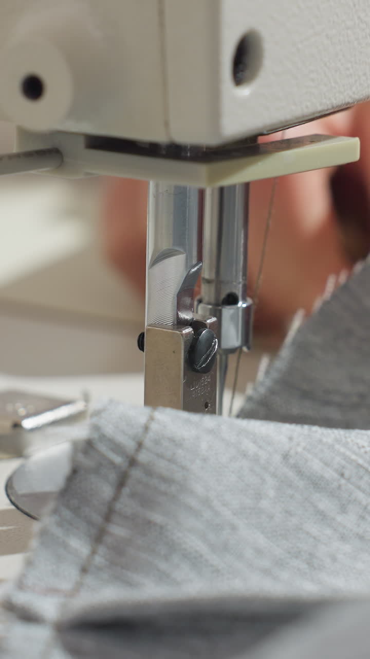 closeup sewing machine needle stitching grey fabric, hands guiding seam under presser foot in bright studio light, precision topstitching on durable knit, focused workshop environment, garment