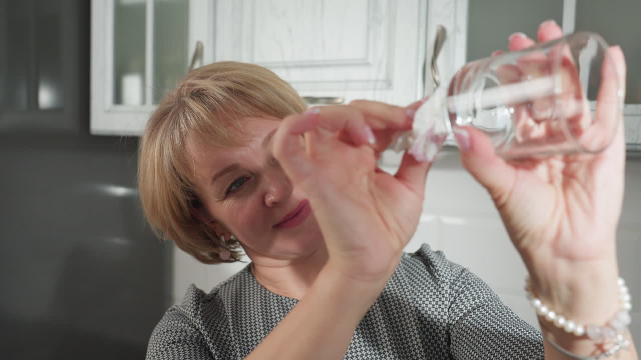 Smiling woman holding transparent glass cup carefully cleans and polishes it with tissue paper, ensuring it is spotless, wearing patterned dress and bracelets, standing in bright modern kitchen